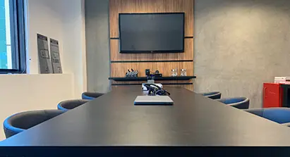 Imagem refletindo uma sala de reunião, com uma mesa preta, três cadeiras de cada lado e na parede uma televisão.