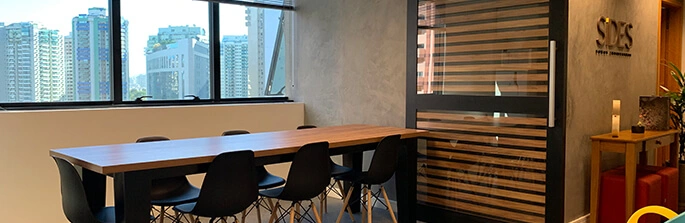 Uma sala para que os funcionários possam socializar ou fazerem refeições, contendo 3 cadeiras de cada lado e uma na ponta de uma mesa preta, ao fundo temos uma janela com vista para prédios.