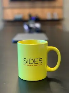 Uma caneca amarela com a logo da agência SIDES sobre a mesa da sala de reunião ao fundo desfocada.